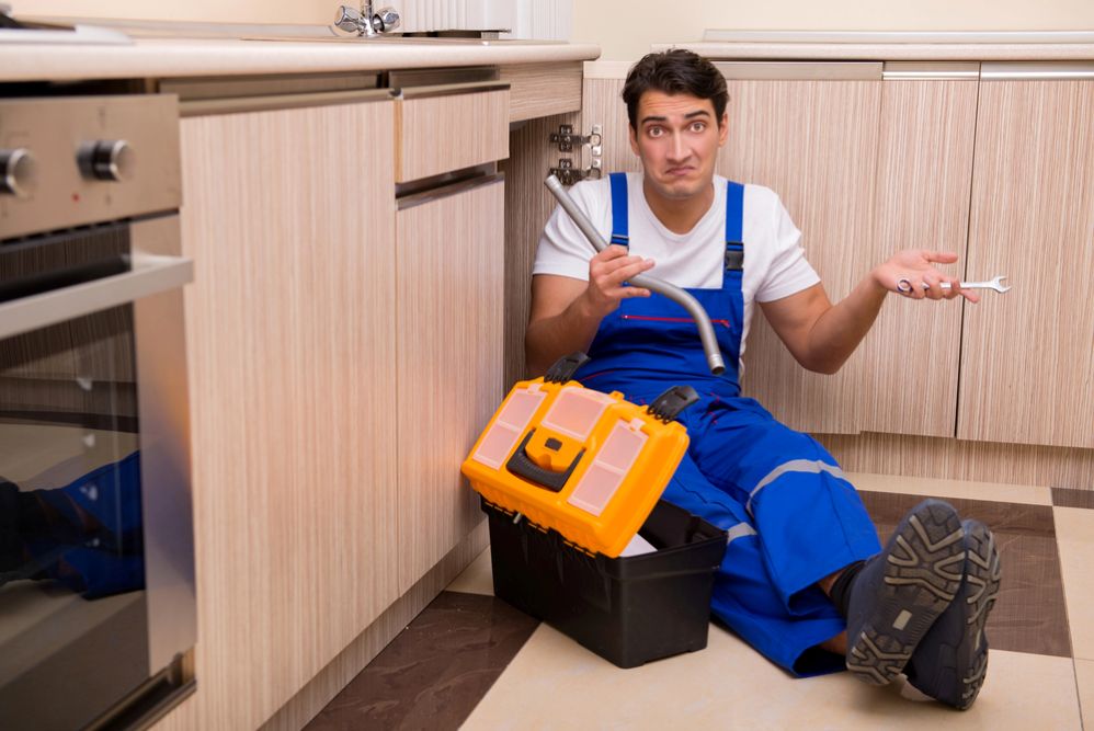 An unlicensed plumber sitting confused under a kitchen sink with broken pipe and tools, showing the risks of hiring unqualified tradies in Belconnen.