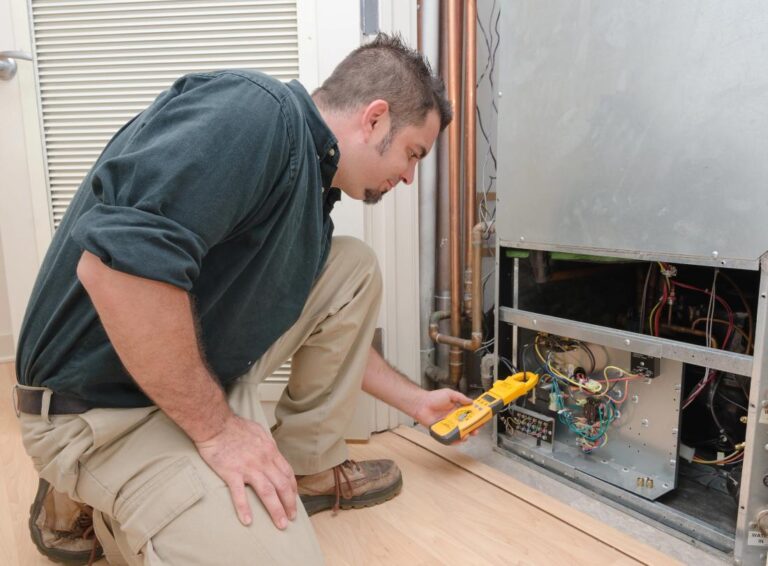 A technician kneeling on a floor using an electrical meter to check wiring inside a home HVAC unit during routine maintenance.