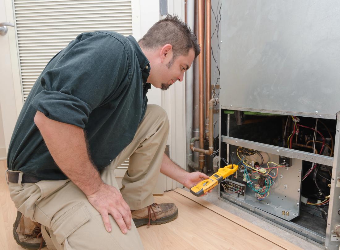 A technician kneeling on a floor using an electrical meter to check wiring inside a home HVAC unit during routine maintenance.